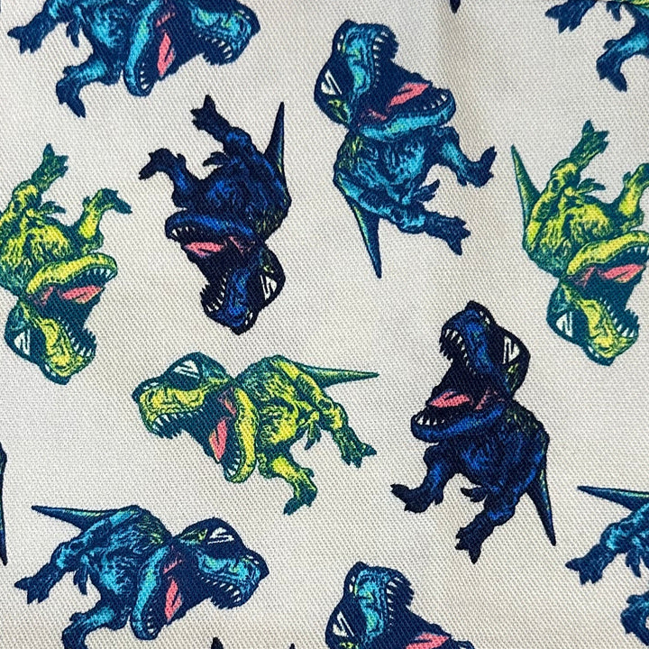 A white fabric head covering with an all-over dinosaur print in blue, green, and yellow, laid flat on a beige textured surface. A black brand tag reading 'SUNSHINE CAP' is visible on the right side.